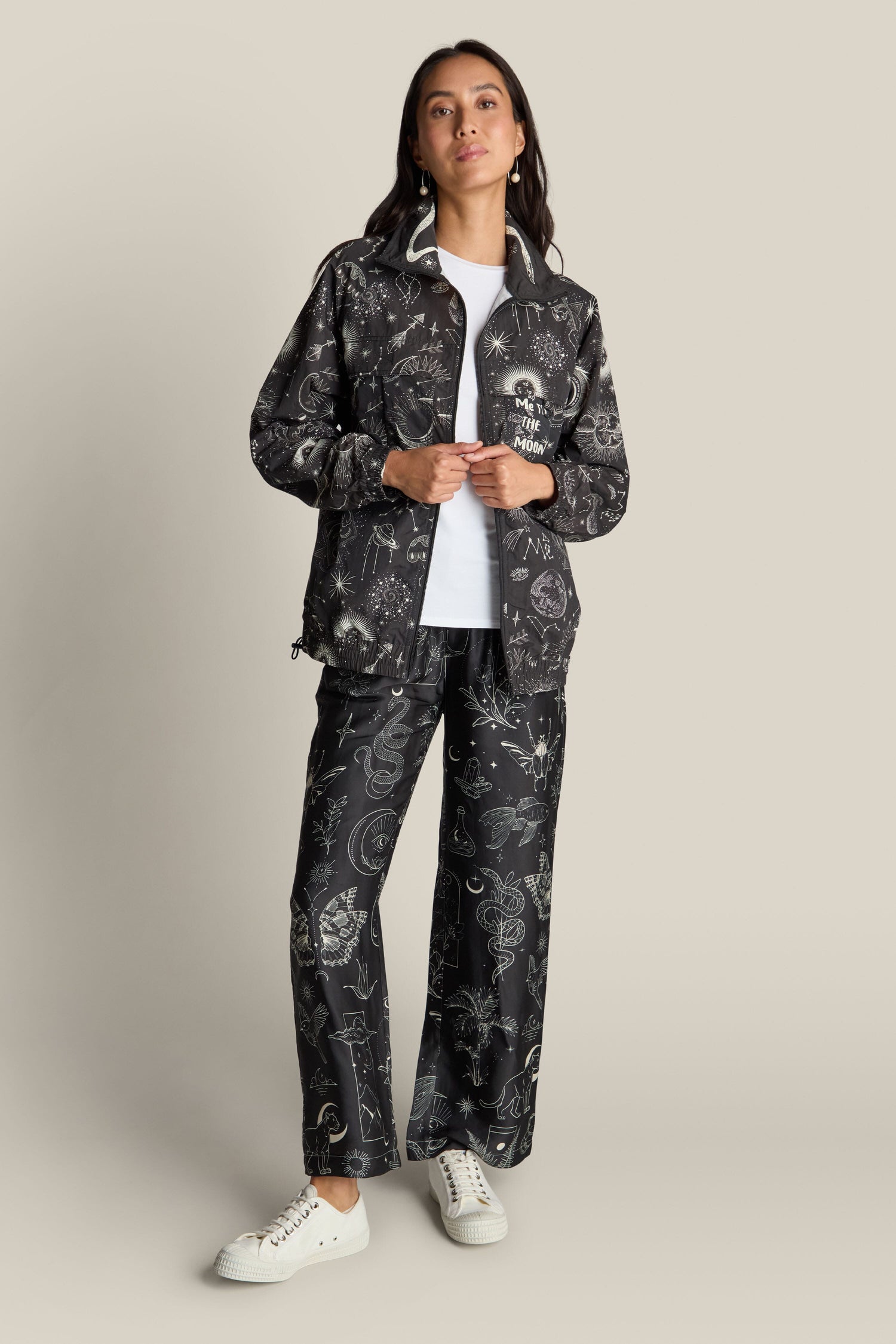 A person stands against a plain background wearing a matching Jolene Track Jacket and pants, adorned with a captivating constellation print. They are paired with a white shirt and white sneakers.