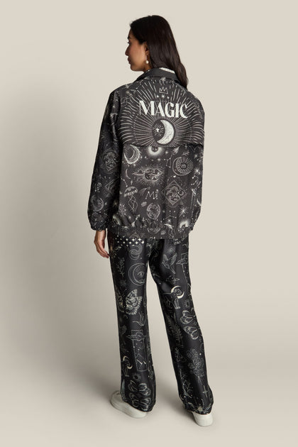 A person facing away, wearing a black Jolene Track Jacket adorned with mystical symbols and "MAGIC" written on the back. They are paired with matching black pants featuring constellation print designs.