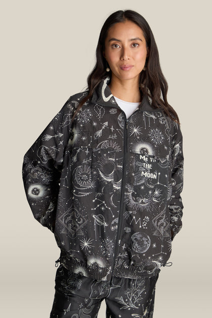 A person donning a Jolene Track Jacket adorned with a dark, celestial-themed constellation print showcasing stars, planets, and the words "Take Me to The Moon." They have long, dark hair and are posing against a plain background.