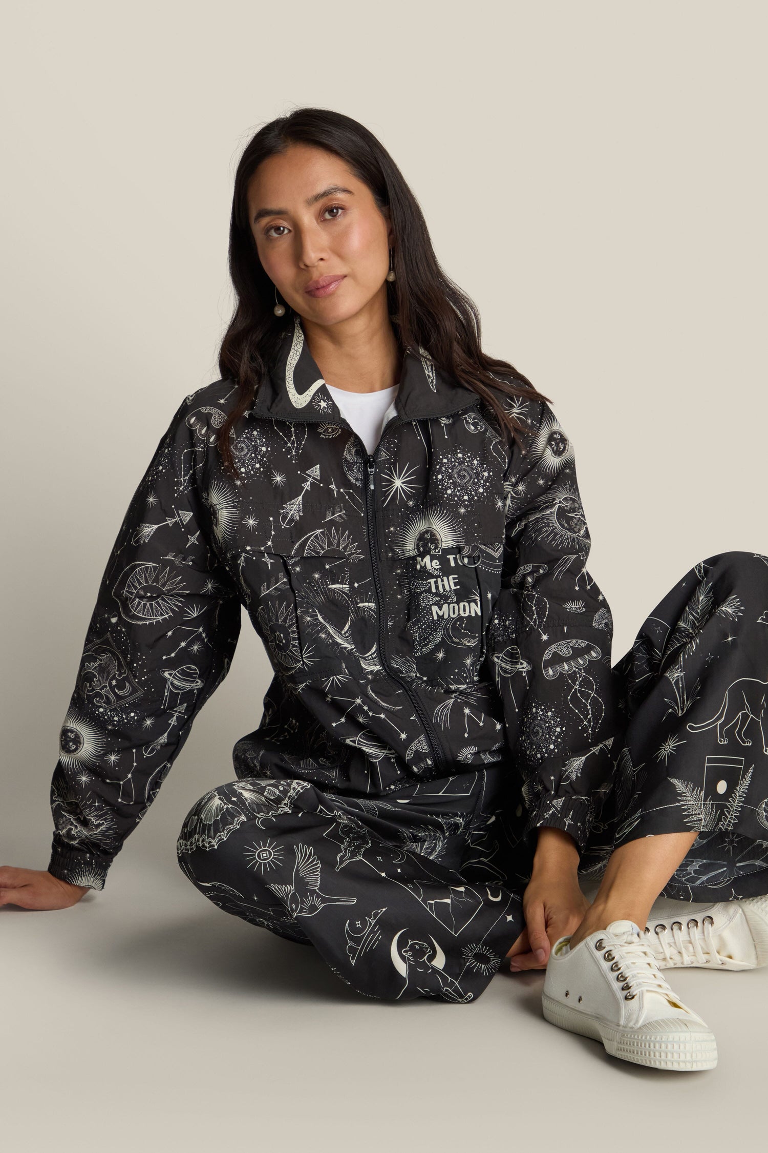 A person sits on the floor wearing the Jolene Track Jacket, which features a black fabric adorned with white whimsical patterns resembling a constellation print, paired with white sneakers against a neutral background.