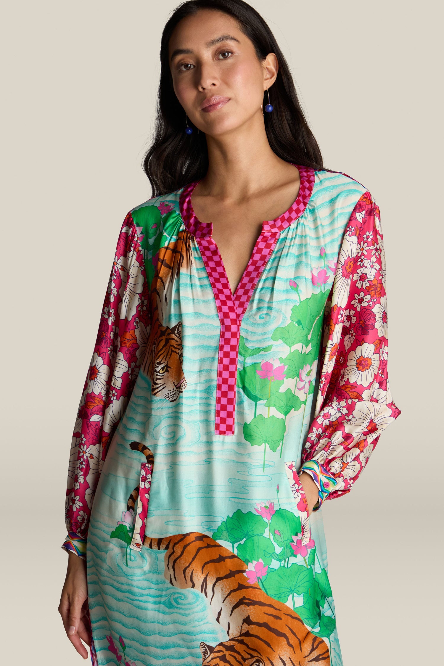 A woman stands against a plain background, showcasing the Twisted Love Hanna Placket Dress, adorned with a vibrant floral design, tiger illustration, long sleeves, and a checkered neckline.