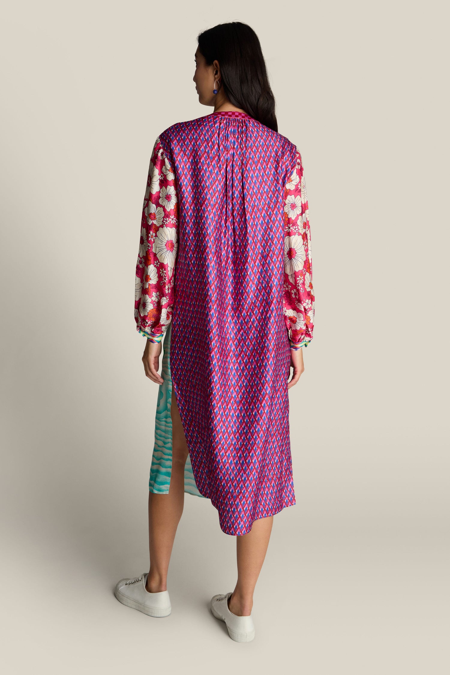 A woman stands wearing a vibrant Twisted Love Hanna Placket Dress, featuring patterned sleeves. The dress has a side slit revealing her white sneakers, and her hair is down, adding an elegant touch to the floral design.