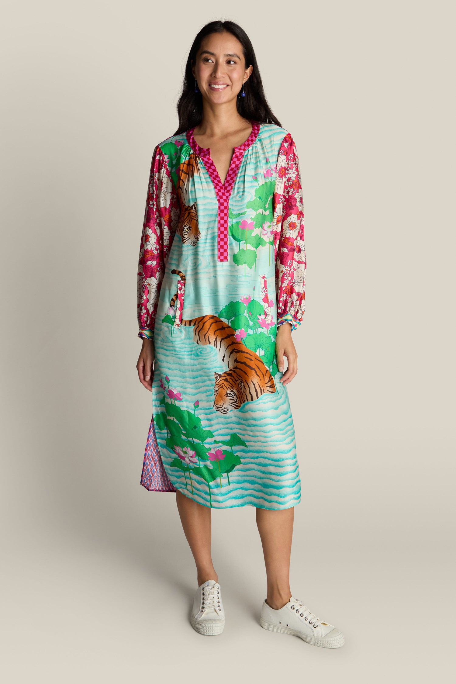 A woman in a vibrant Twisted Love Hanna Placket Dress, adorned with tiger illustrations and floral designs, stands against a plain background. She wears white sneakers and is smiling.