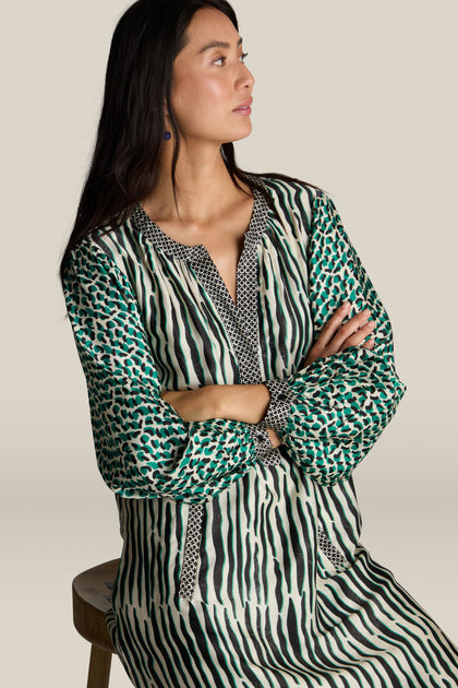 A woman in a Tropical Hanna Placket Dress by ME369 sits on a stool with her arms crossed, looking to her left against a plain beige background.