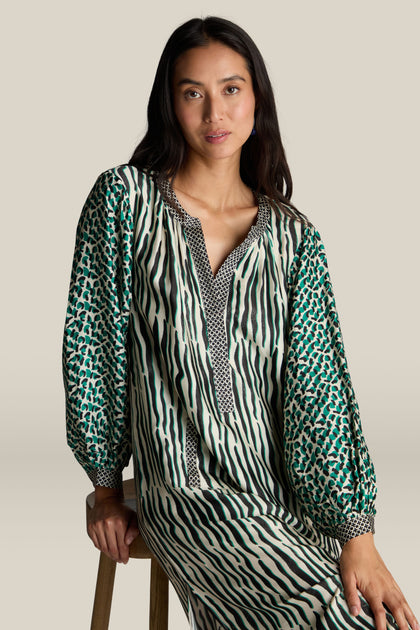 A woman with long dark hair wearing the Tropical Hanna Placket Dress sits on a wooden stool against a plain background.