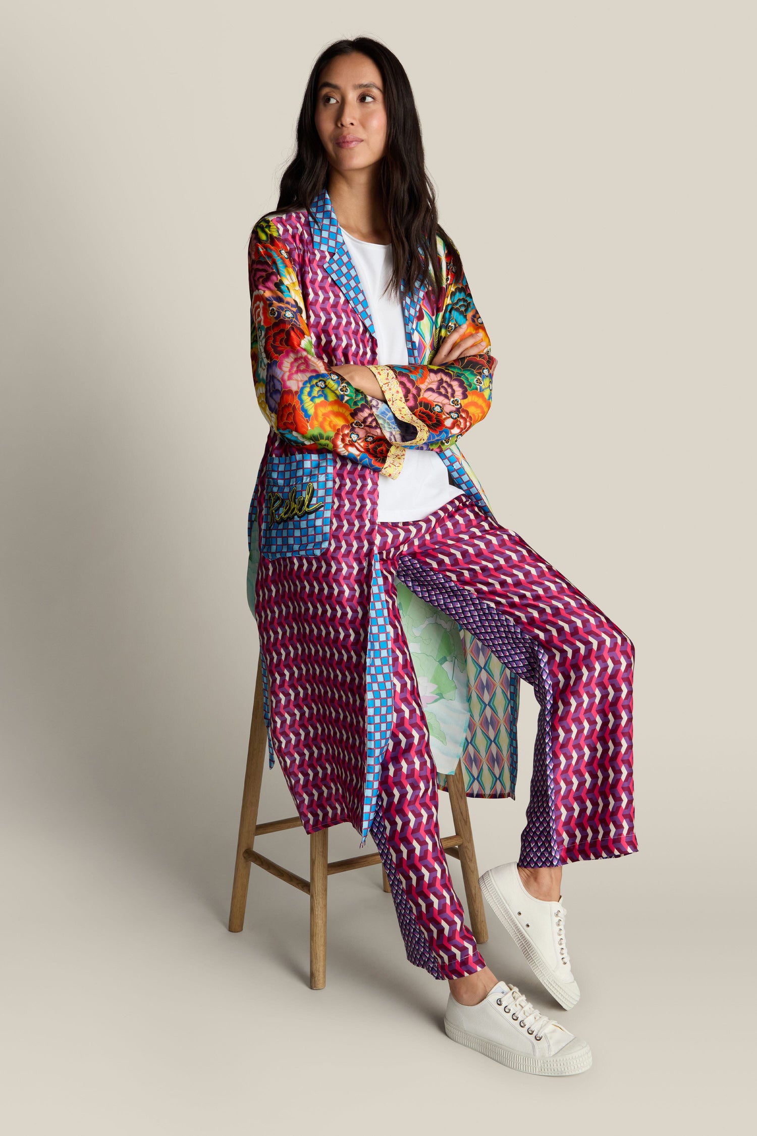 A person sits on a stool wearing the striking Nova Kimono, paired with a vibrant, patterned outfit showcasing a delicate tiger illustration and complemented by white sneakers. They look forward with arms crossed against a plain beige background.