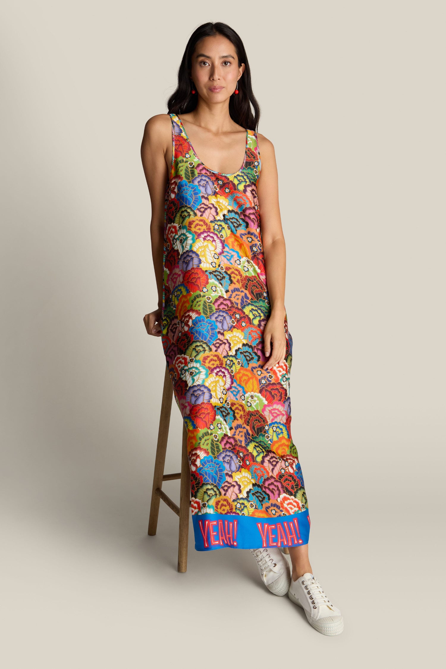 A person wearing the Allison Dress, a colorful, patterned maxi dress featuring a vibrant floral design and the word "YEAH!" written near the hem, stands next to a wooden stool against a neutral background.