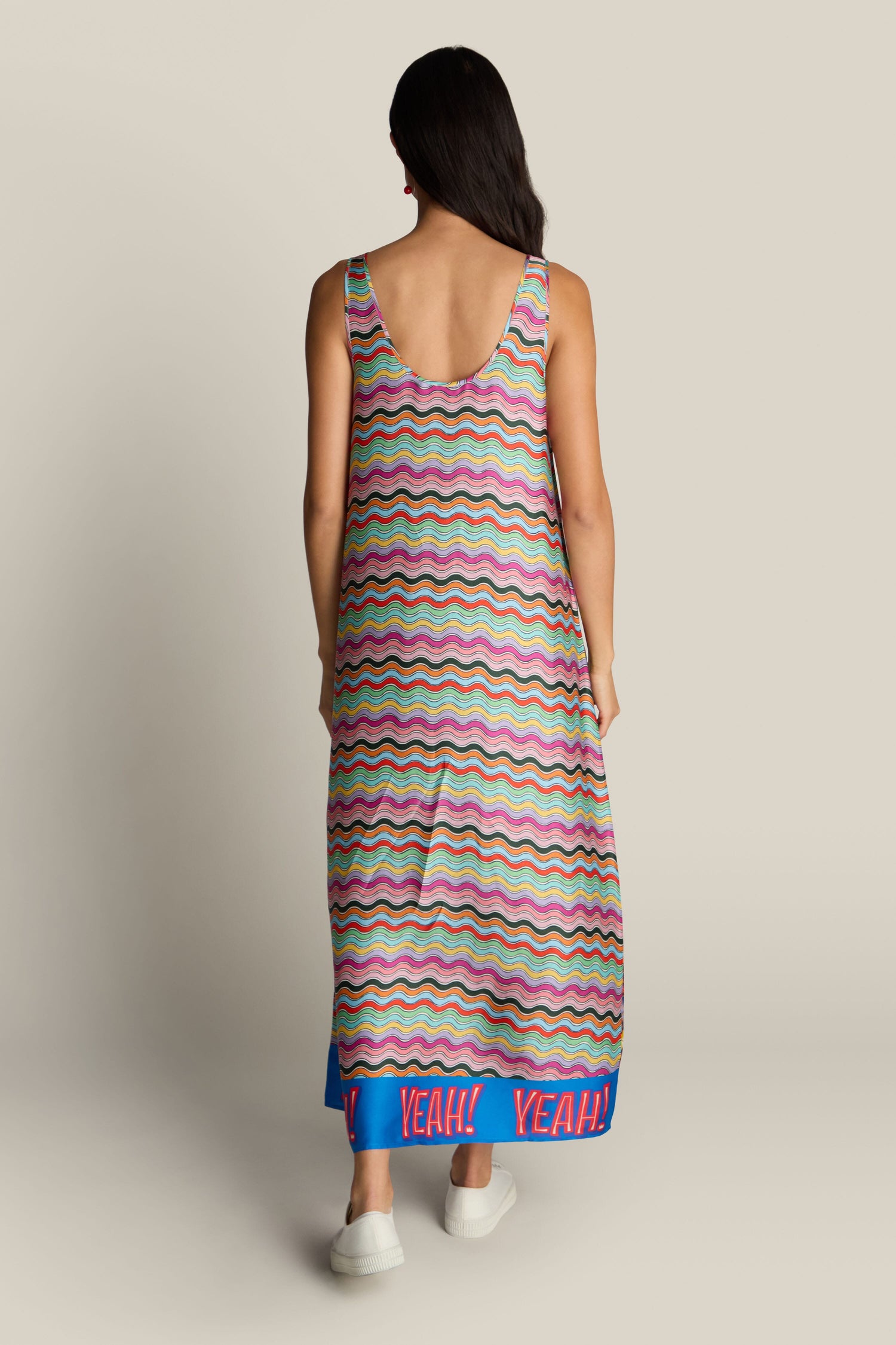 A person with long hair, seen from the back, wearing a colorful Allison Dress featuring a wavy-patterned maxi design and "YEAH!" printed along the blue hemline, complemented by white shoes.