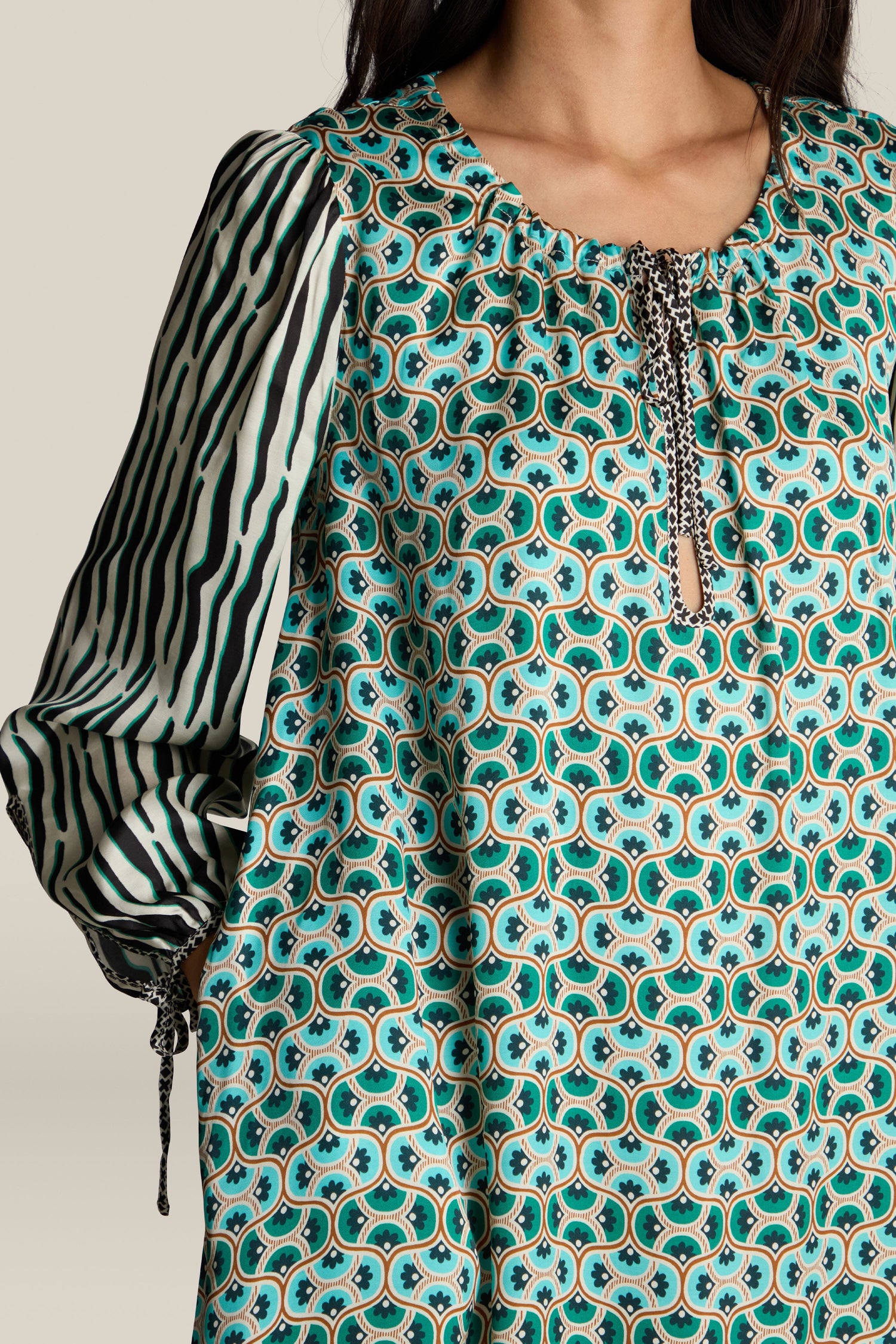 A person wearing the Polette Dress, featuring a green and blue patterned design with black and white striped sleeves and a tie detail at the neckline. The ME369 dress showcases a unique digital print. Only part of the person's face is visible.