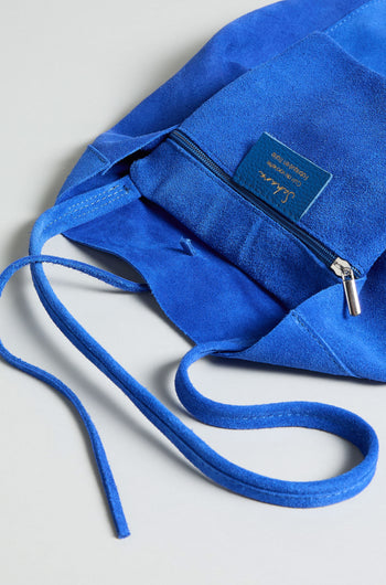 Close-up of the Soft Suede Tote Bag in royal blue, featuring a zippered pocket, visible brand label, and matching suede drawstrings on a light gray surface—an elegant minimalist tote for effortless style.