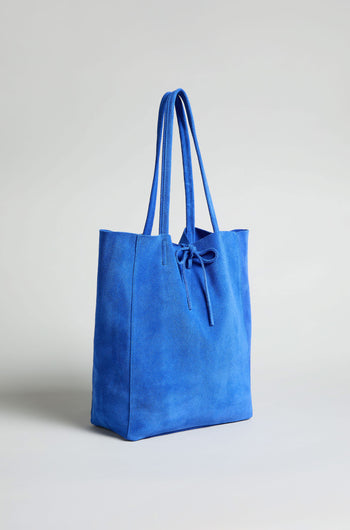 The Soft Suede Tote Bag, in blue with long handles and a front tie detail, is displayed against a plain light background.