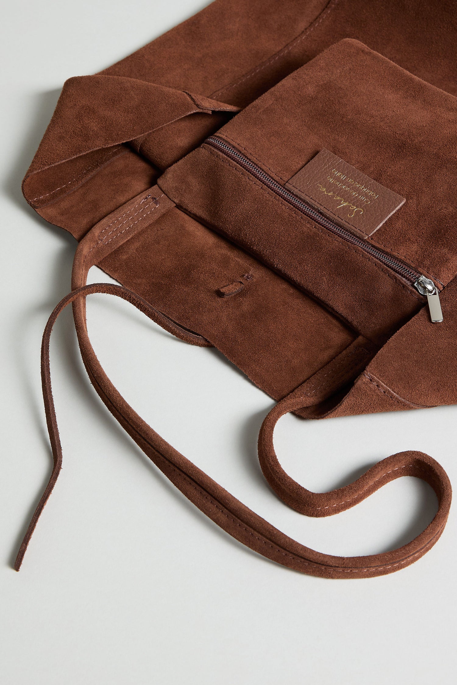The Soft Suede Tote Bag, featuring brown suede, visible stitching, an open zipper pocket, and a long strap, rests on a flat surface—ideal for anyone who loves a minimalist tote with understated style.