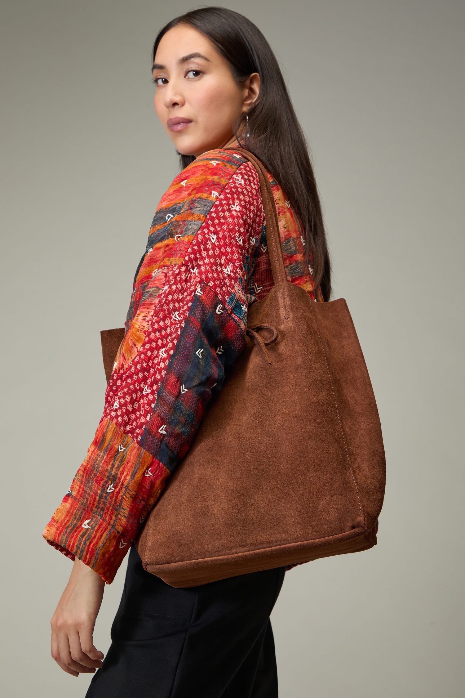 A woman with long dark hair wears a colorful patterned jacket and carries the Soft Suede Tote Bag, a large brown luxury accessory, over her shoulder while facing slightly backward.