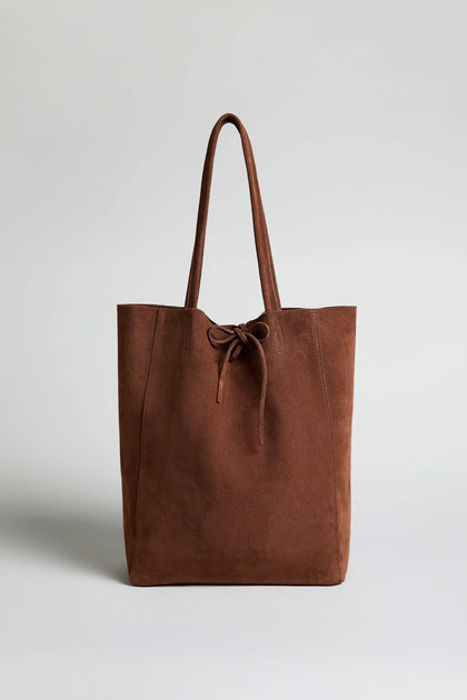 The Soft Suede Tote Bag, a brown minimalist tote with two handles and a front tie detail, is shown upright against a plain light background.