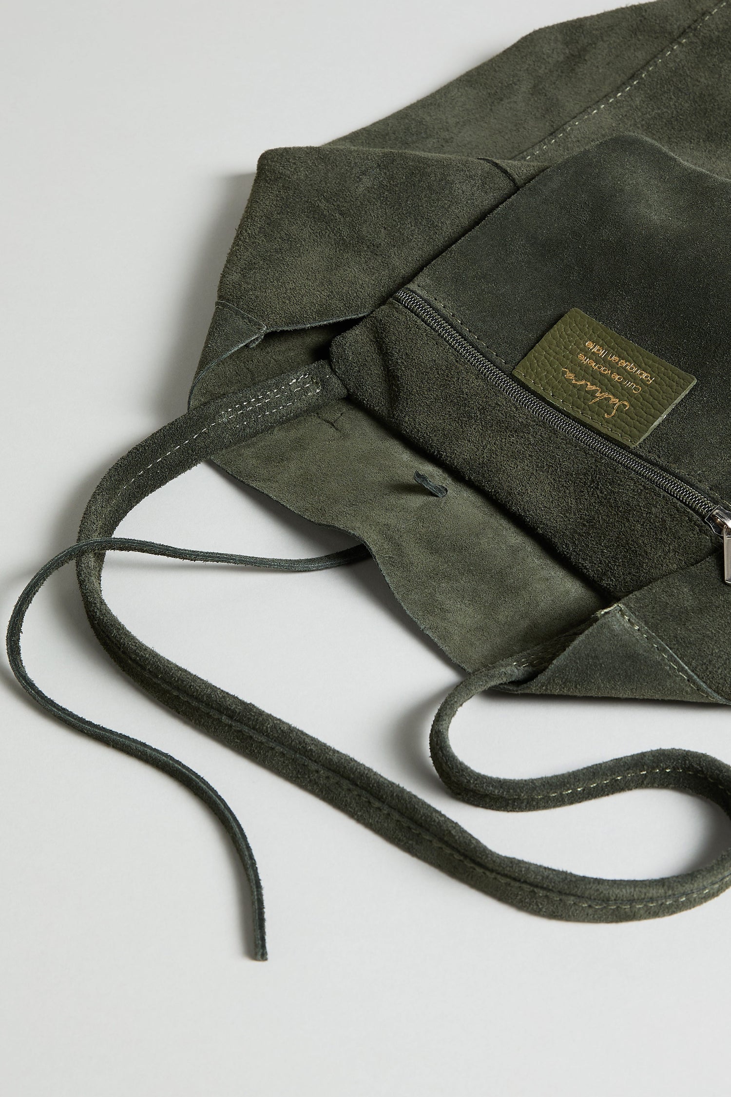 The Soft Suede Tote Bag in olive green features a drawstring closure, front zipper pocket, and a small rectangular label. This minimalist suede tote is displayed flat on a light background.