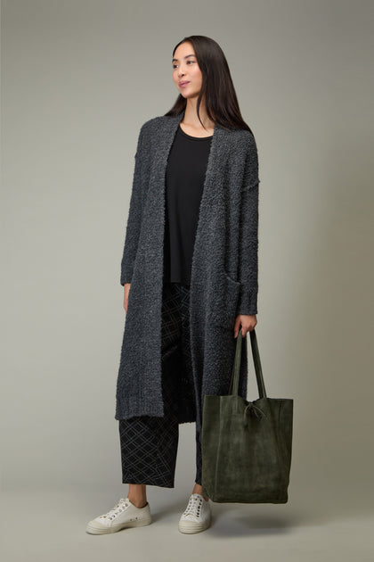 Woman stands against a neutral background wearing a long gray cardigan, black top, patterned pants, white sneakers, and holding the Soft Suede Tote Bag in green.