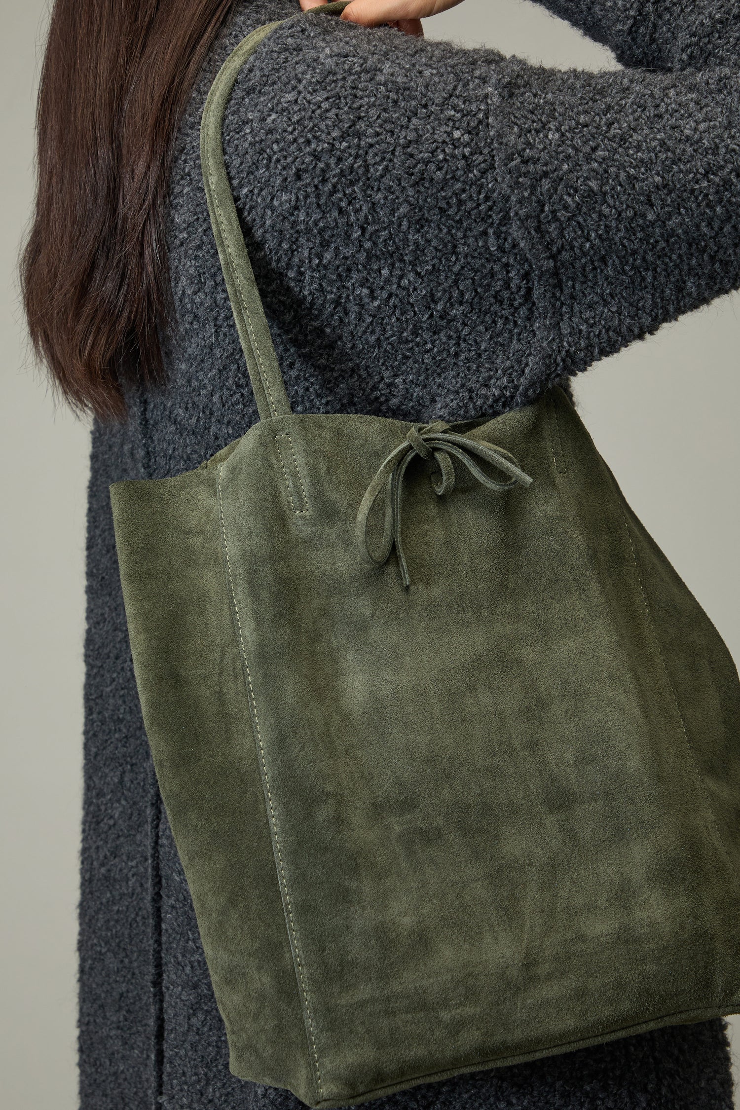 A person in a textured dark gray coat carries the Soft Suede Tote Bag, a large olive green luxury tote with a simple tie closure, over their shoulder.
