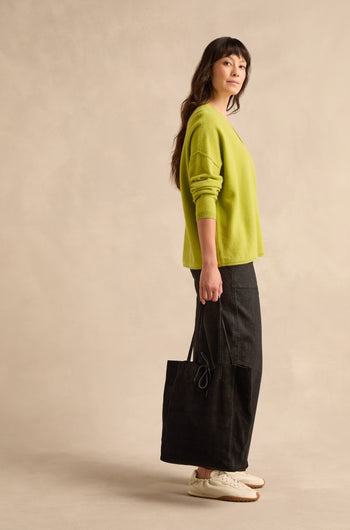 A woman stands sideways against a neutral background, dressed in a lime green sweater, long black skirt, and white sneakers, holding the Soft Suede Black Tote Bag on her shoulder.