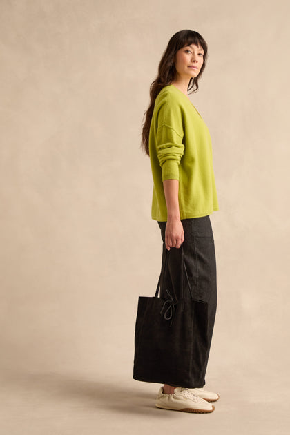 A woman stands sideways against a neutral background, dressed in a lime green sweater, long black skirt, and white sneakers, holding the Soft Suede Black Tote Bag on her shoulder.