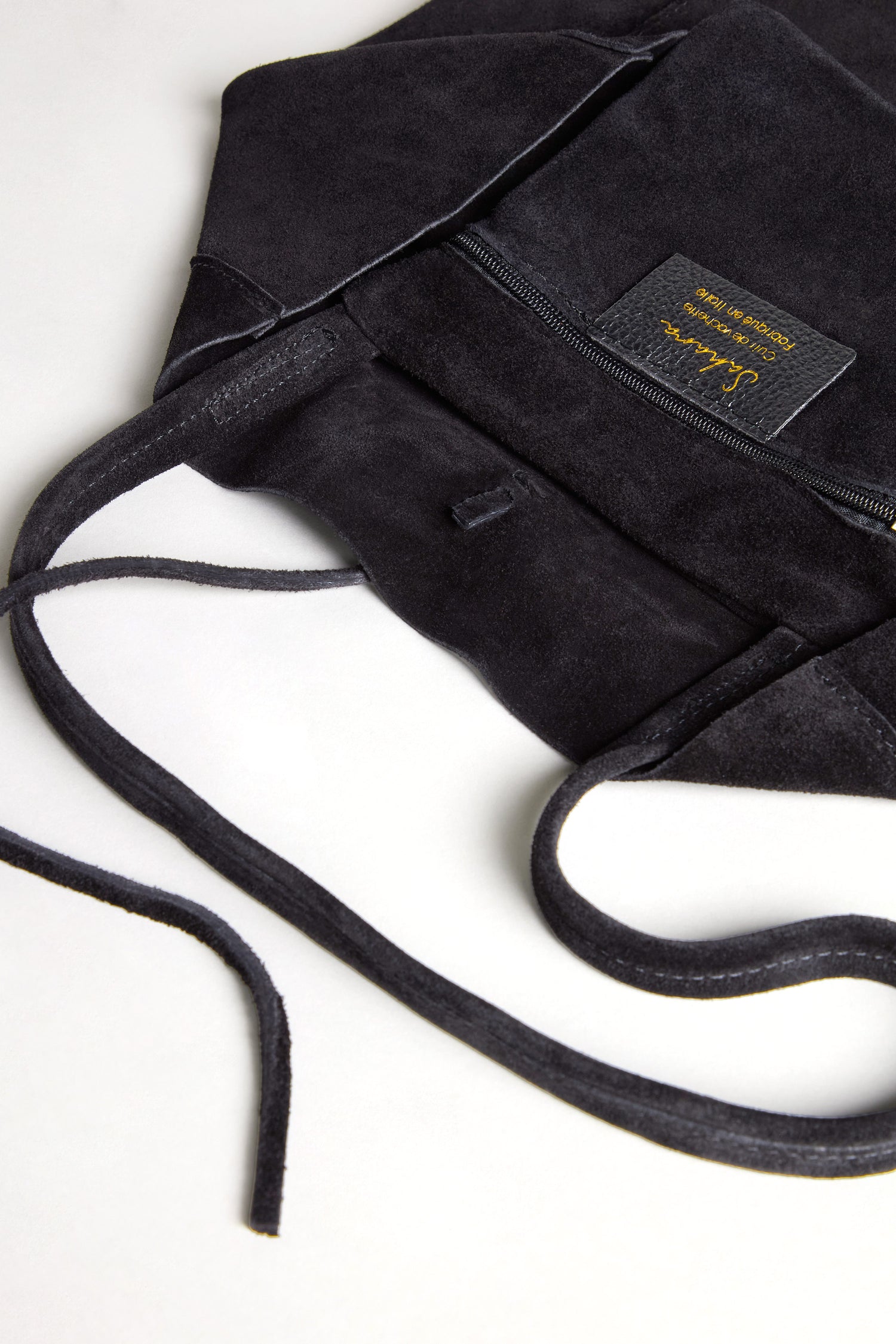 The Soft Suede Black Tote Bag features a luxurious black suede exterior, a front zipper pocket, and gold lettering on its label. Laid open on a white surface, this spacious shoulder bag seamlessly merges the elegance of suede with minimalist style.