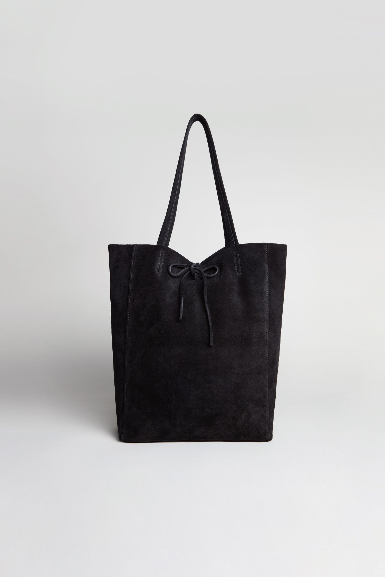 The Black Soft Suede Tote Bag, shown against a plain light background, features a spacious design with two handles and a charming bow detail on the front.