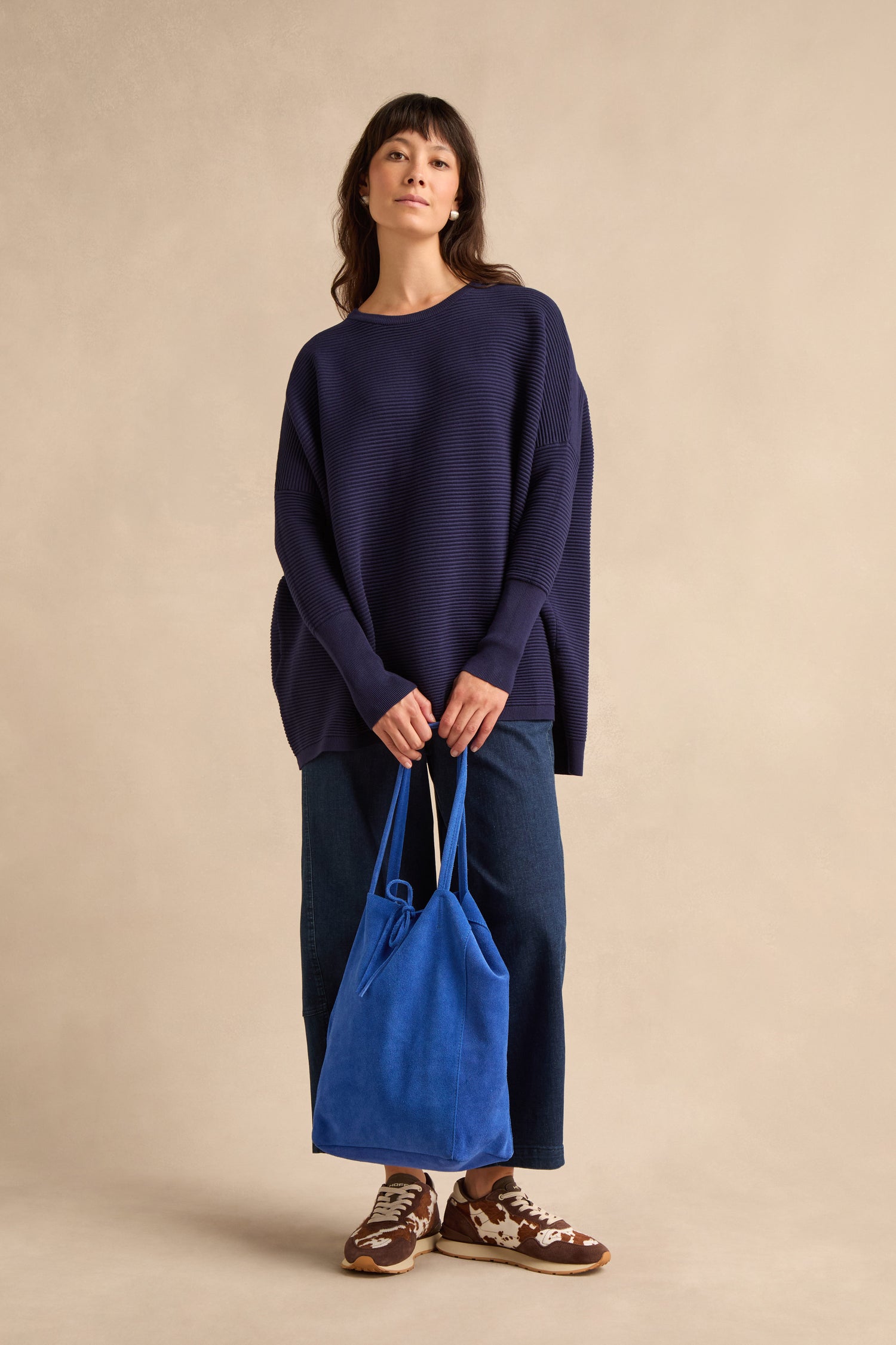 A woman stands against a beige background, wearing a loose navy sweater, wide-leg jeans, brown sneakers, and holding the Blue Soft Suede Tote Bag—a minimalist handbag in bright blue.