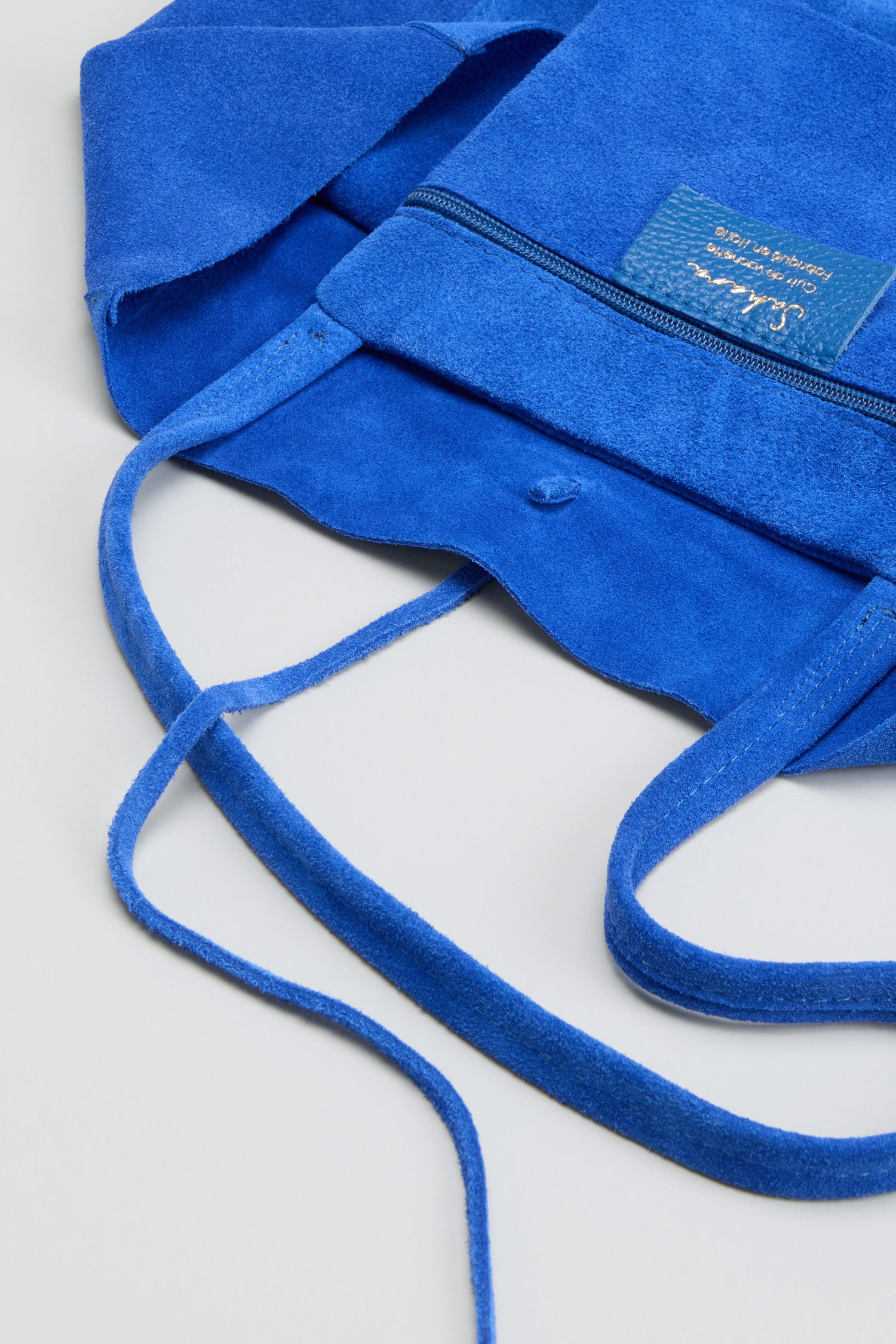 Introducing the Blue Soft Suede Tote Bag—an elegant, minimalist handbag featuring an open top design, front zipper pocket, slim straps, and a visible interior fabric tag. Presented on a light surface, it’s crafted for effortless style.
