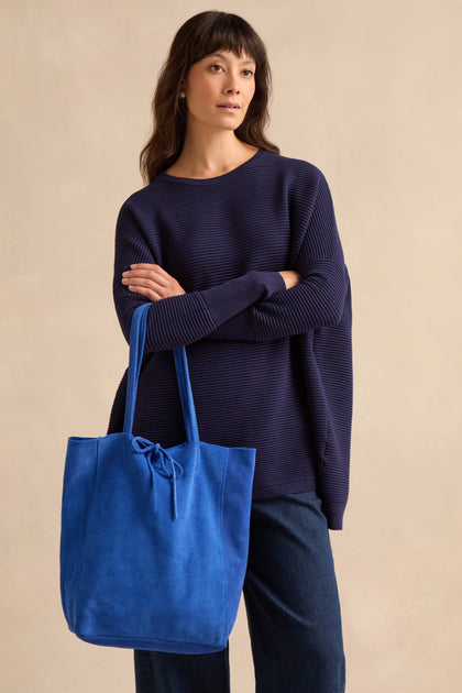A person in a navy sweater and jeans stands against a neutral background, holding the Blue Soft Suede Tote Bag—a large tote with tied handles—in their hand.