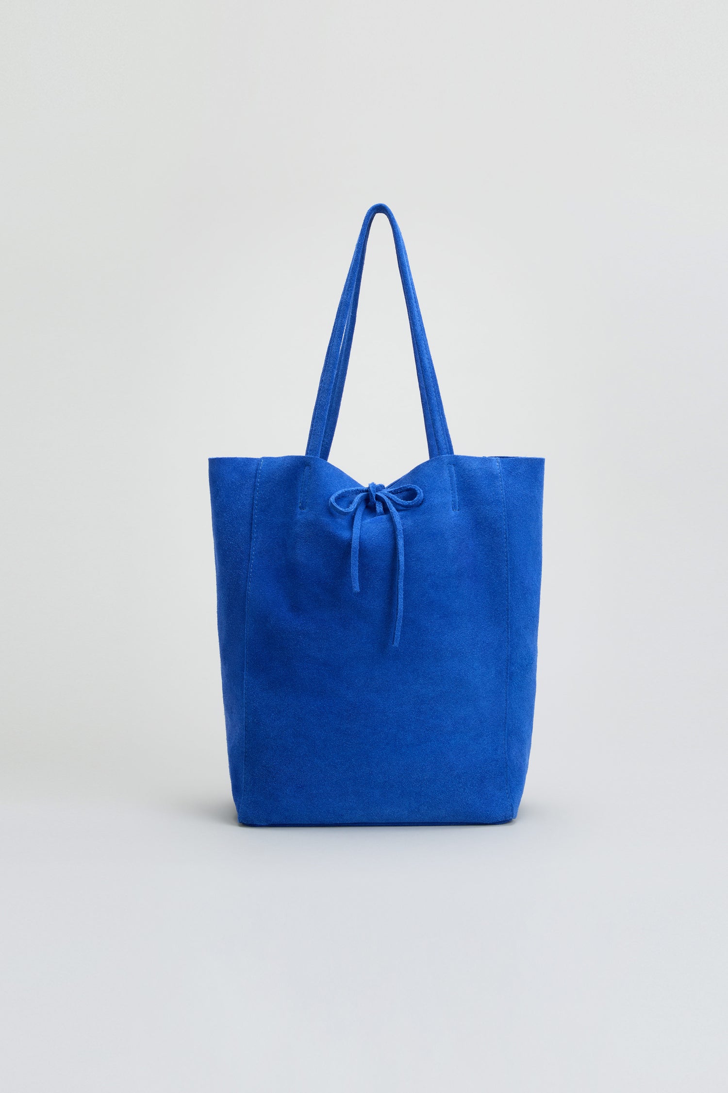 The Blue Soft Suede Tote Bag, featuring a vibrant blue suede exterior, two handles, and a drawstring closure, is showcased against a simple light background.