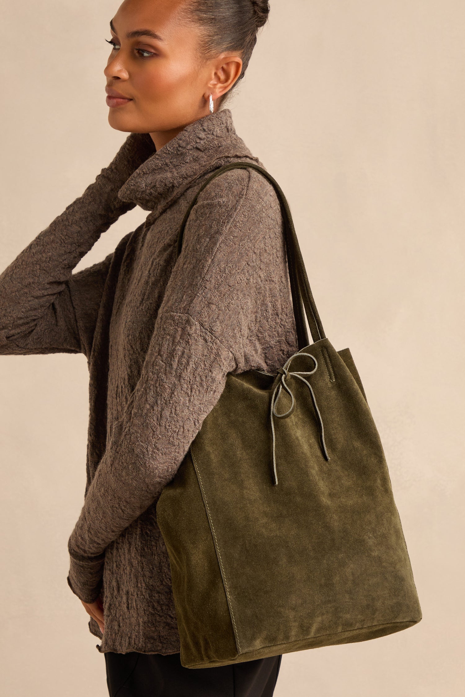 A woman stands against a neutral background wearing a textured brown sweater and carrying the Soft Suede Khaki Tote Bag—spacious and olive green with a bow detail—on her shoulder.