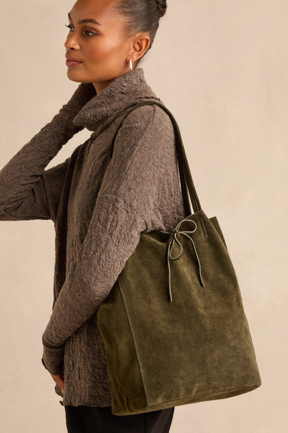 A woman stands against a neutral background wearing a textured brown sweater and carrying the Soft Suede Khaki Tote Bag—spacious and olive green with a bow detail—on her shoulder.