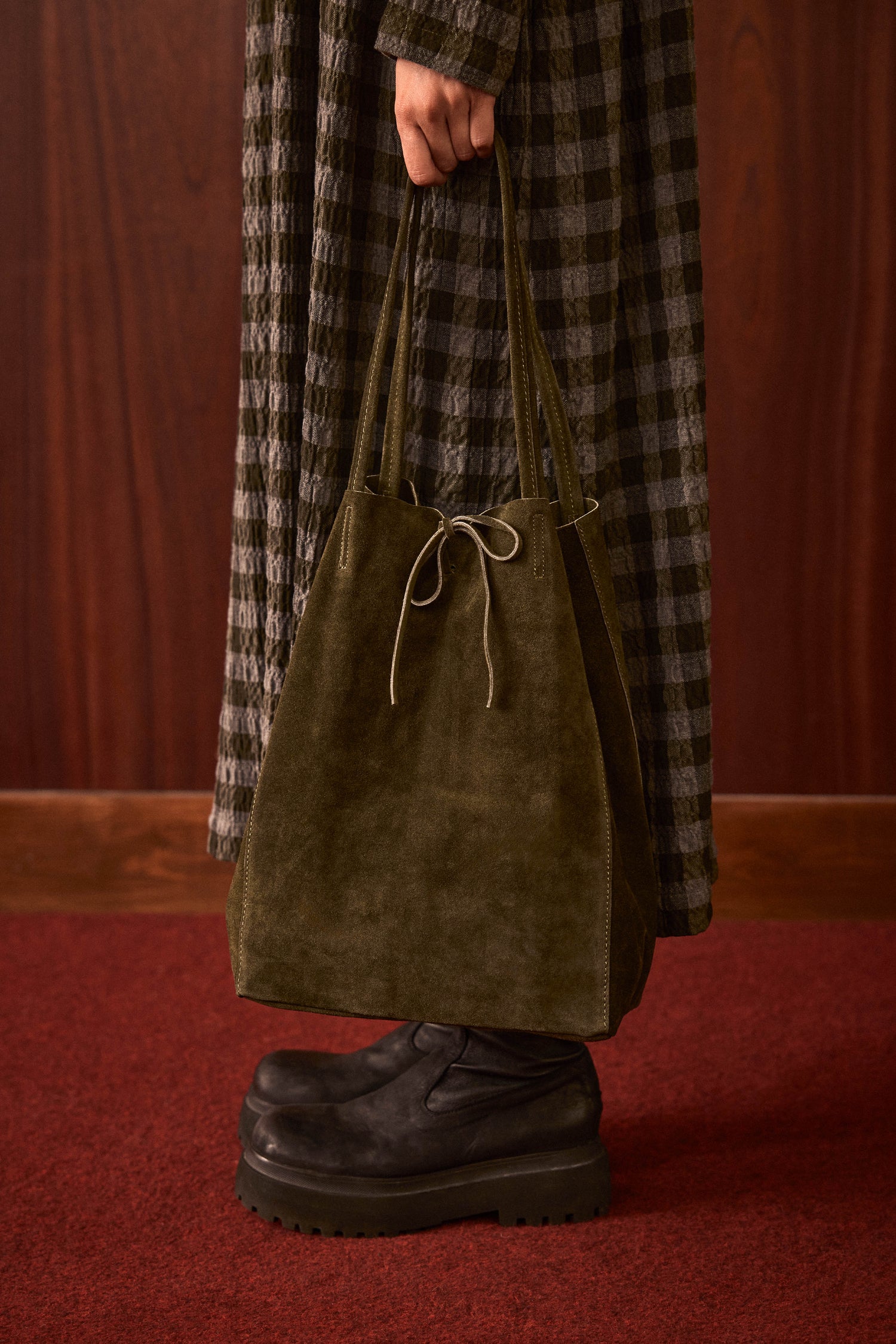 Standing on a red carpet, the person wears a plaid coat and black platform shoes, holding the Khaki Soft Suede Tote Bag by its handles.