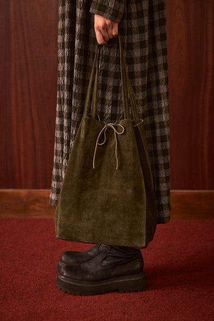 Standing on a red carpet, the person wears a plaid coat and black platform shoes, holding the Khaki Soft Suede Tote Bag by its handles.