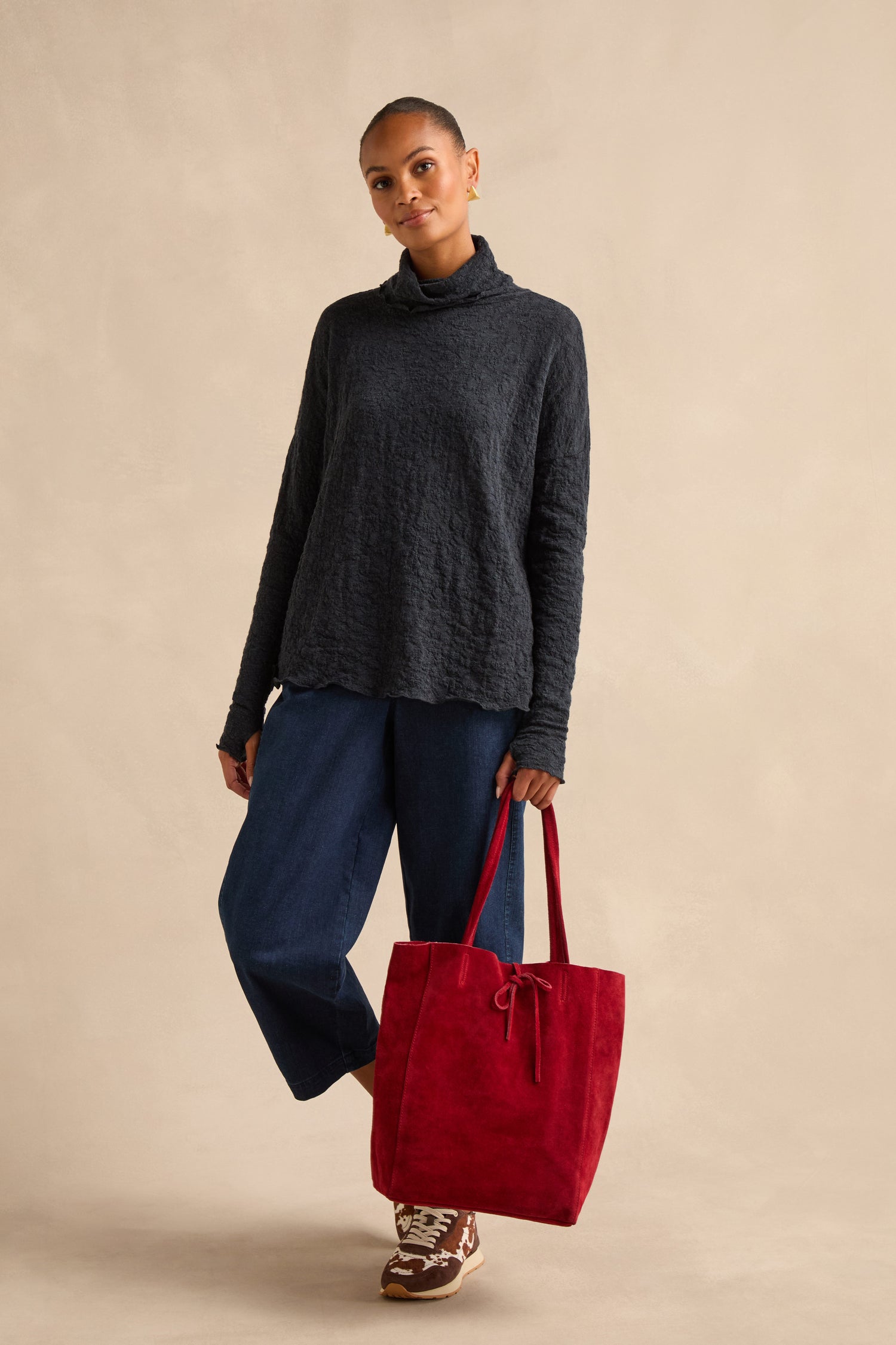 A person stands against a beige background wearing a black sweater, blue jeans, brown sneakers, and holding the Soft Suede Poppy Tote Bag with a spacious interior.