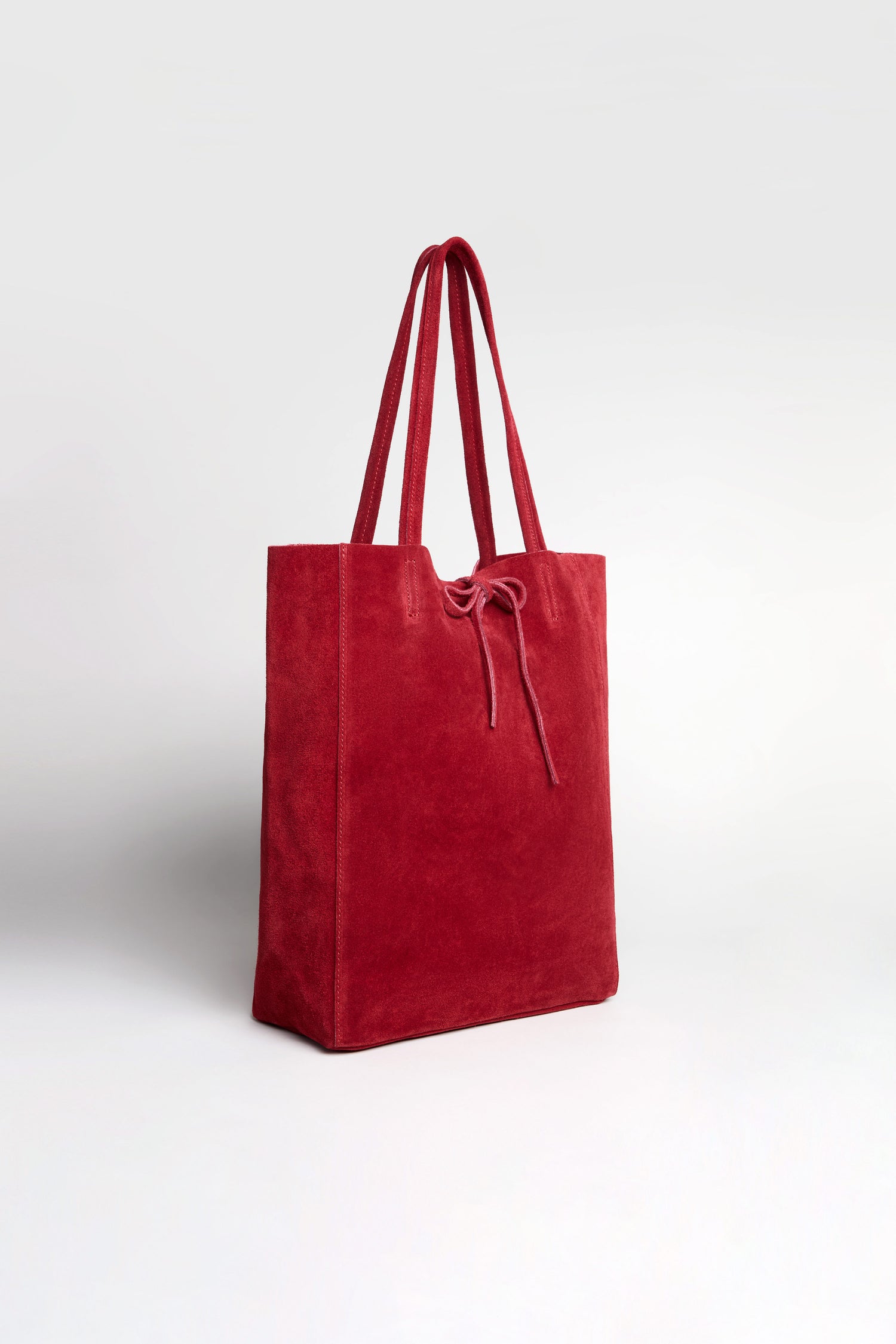 The Soft Suede Poppy Tote Bag is a red tote crafted from soft suede, designed with two handles and a drawstring closure. Featuring a minimalist aesthetic and a spacious interior, it is showcased against a plain light background.