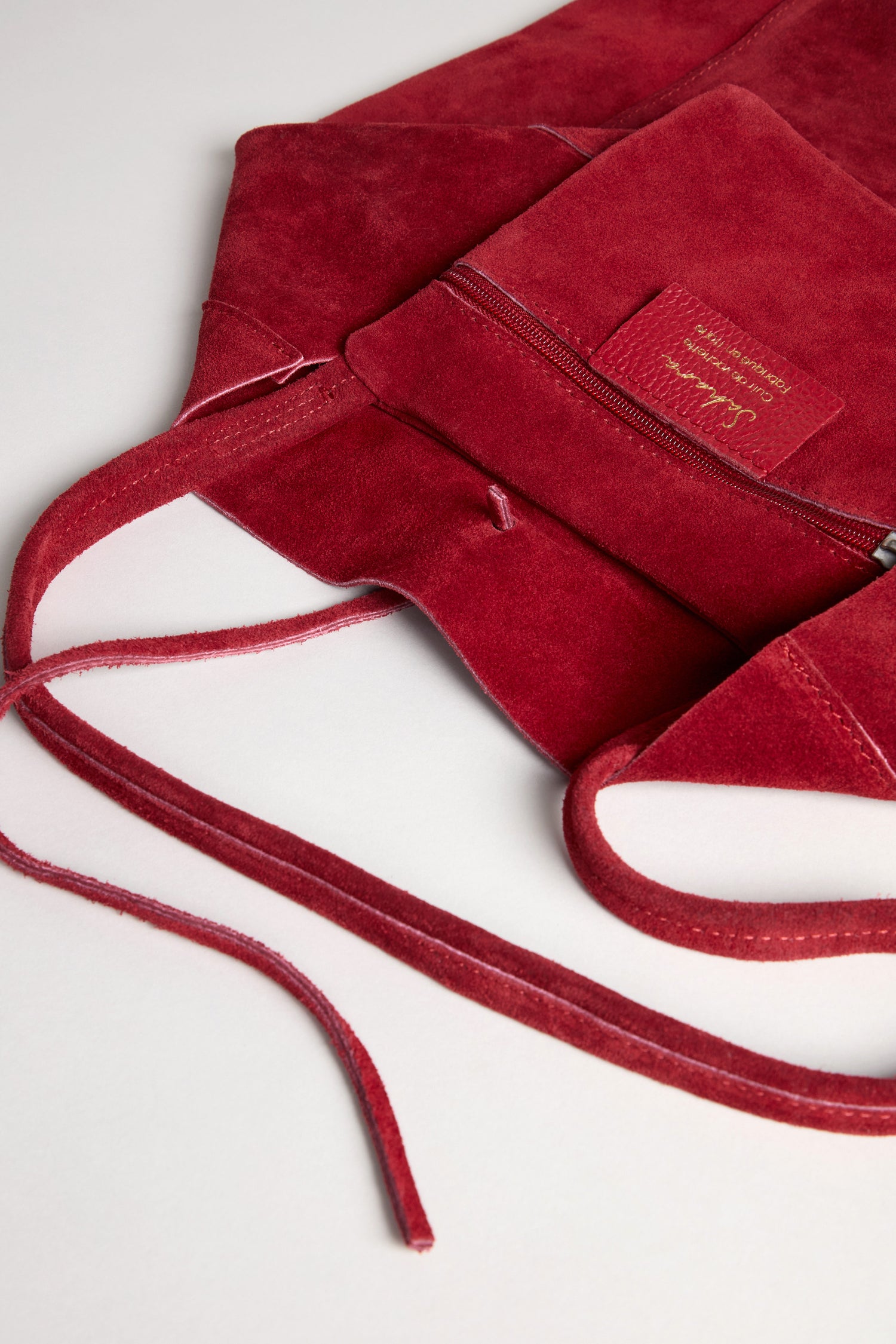 Close-up of the Soft Suede Poppy Tote Bag in red, showcasing its soft suede material, front zipper pocket, stitched label, and long, slender shoulder straps against a light background. The tote’s minimalist design is enhanced by its spacious interior, perfect for carrying everyday essentials.