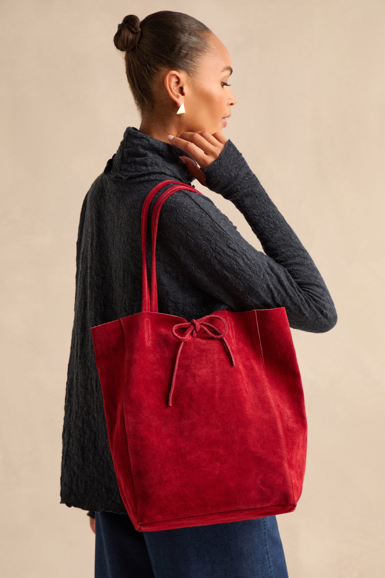 A person in a dark textured top and jeans stands sideways, carrying the Soft Suede Poppy Tote Bag with a tied bow detail over their shoulder. The tote features a spacious interior, perfect for carrying everyday essentials.