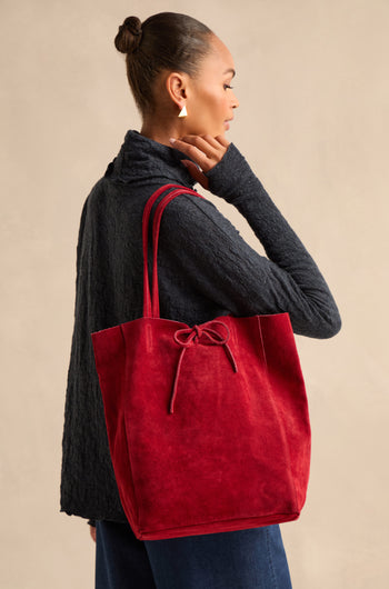 A person in a dark textured top and jeans stands sideways, carrying the Soft Suede Poppy Tote Bag with a tied bow detail over their shoulder. The tote features a spacious interior, perfect for carrying everyday essentials.