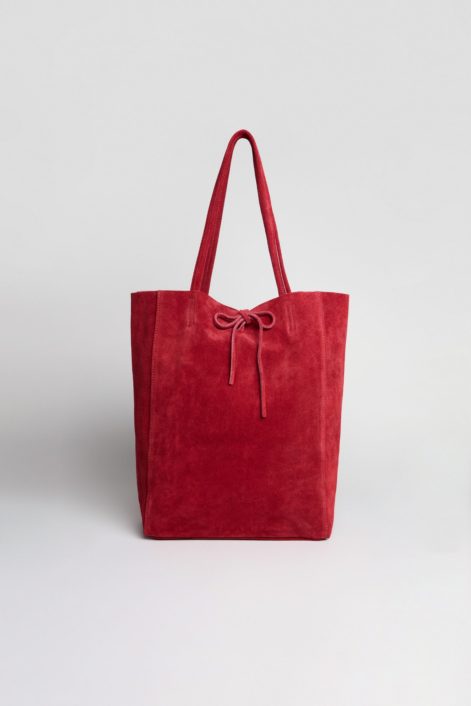 The Soft Suede Poppy Tote Bag is a red minimalist tote, crafted from soft suede, featuring two handles and a drawstring closure. Shown against a plain white background, its spacious interior is ideal for stylishly carrying your daily essentials.