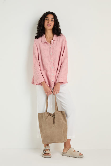 Woman standing against a white wall, wearing a pink button-up shirt, white pants, beige sandals, and holding the Soft Suede Sand Tote Bag.