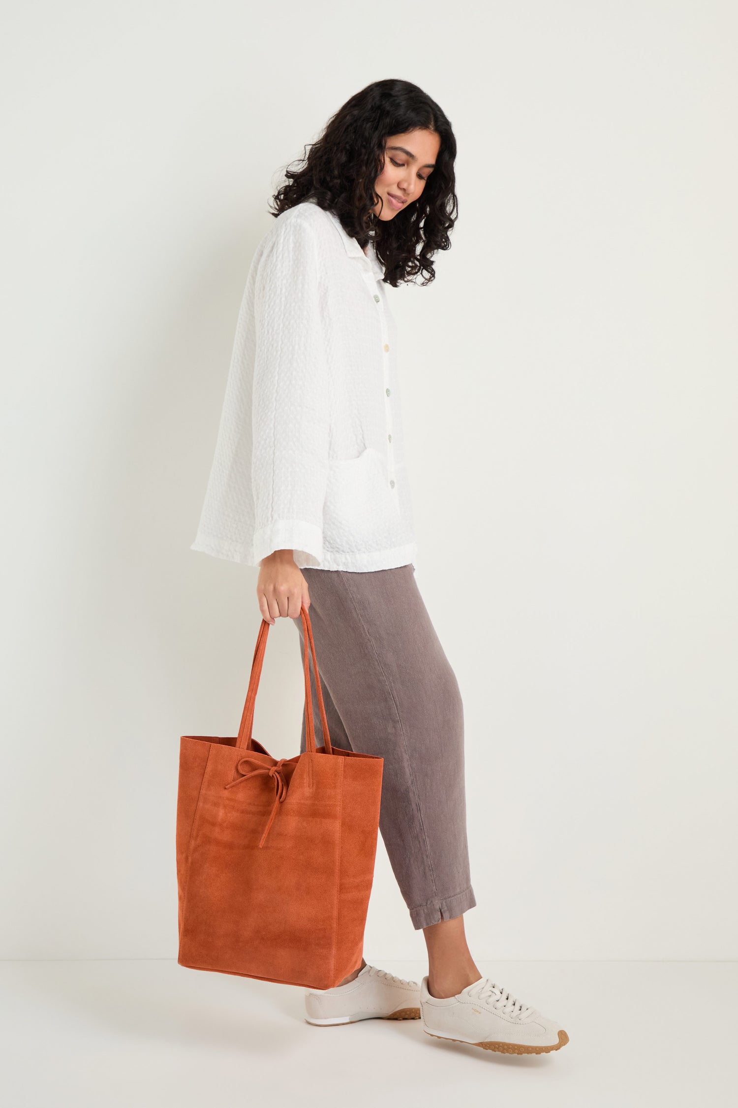 A woman stands against a white wall, dressed in a white button-up shirt, gray pants, white sneakers, and holding the Soft Suede Tangerine Tote Bag—an accessory that perfectly complements her minimalist style.