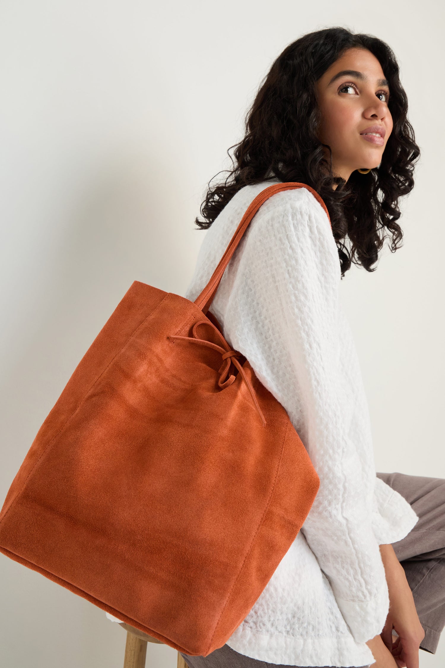 A woman with curly hair sits on a stool against a plain background, wearing a white textured top and holding the Soft Suede Tangerine Tote Bag, known for its spacious interior.
