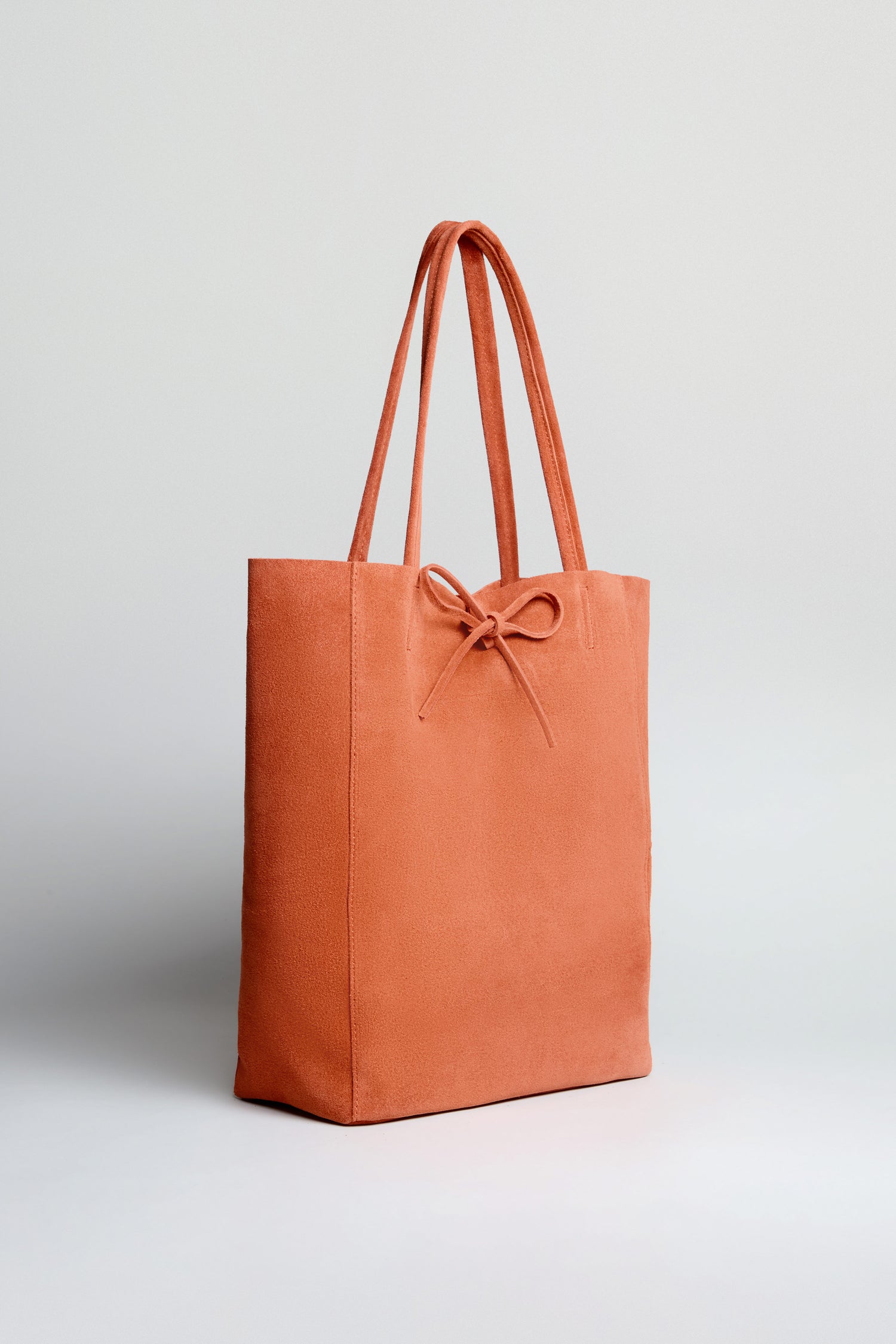 The Soft Suede Tangerine Tote Bag is a rust-colored minimalist tote with two handles, a front bow, and a spacious interior, shown against a plain light background.