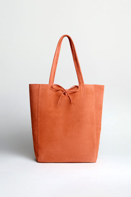 The Soft Suede Tangerine Tote Bag features a muted orange hue, spacious interior, two handles, and a small front bow, shown against a plain white background.
