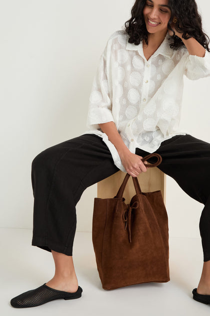 A woman sits on a wooden block, wearing a white textured blouse, black pants, black shoes, and holding the Soft Suede Chocolate Tote Bag.