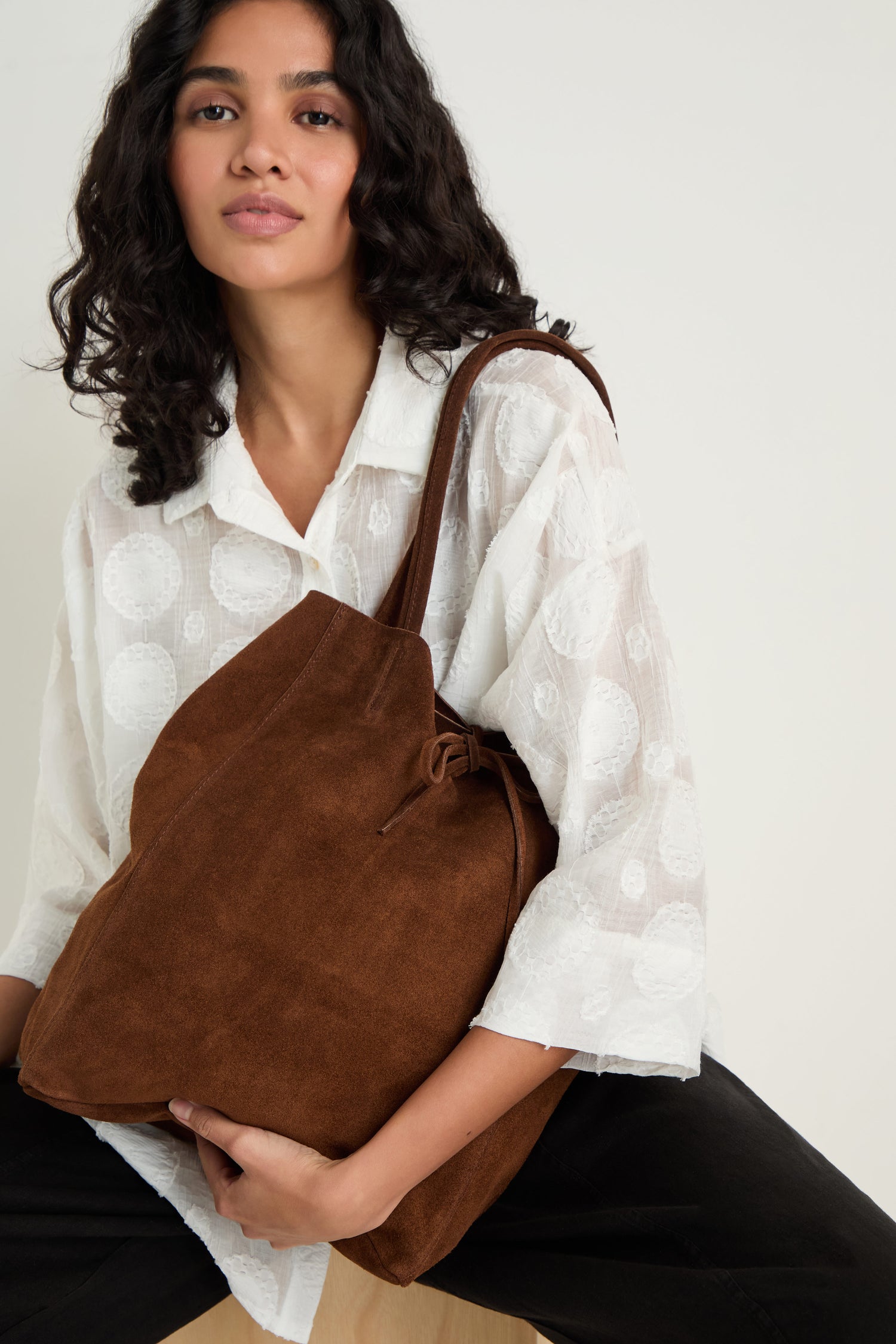 A woman in a white patterned blouse and black pants sits against a plain background, holding the Soft Suede Chocolate Tote Bag.