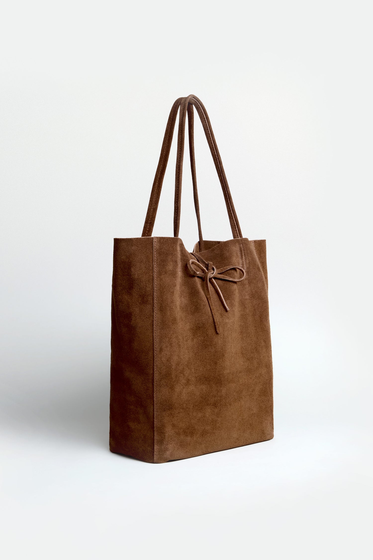 The Soft Suede Chocolate Tote Bag features long handles and a simple bow detail on the front, showcased in a minimalist style against a plain light background.