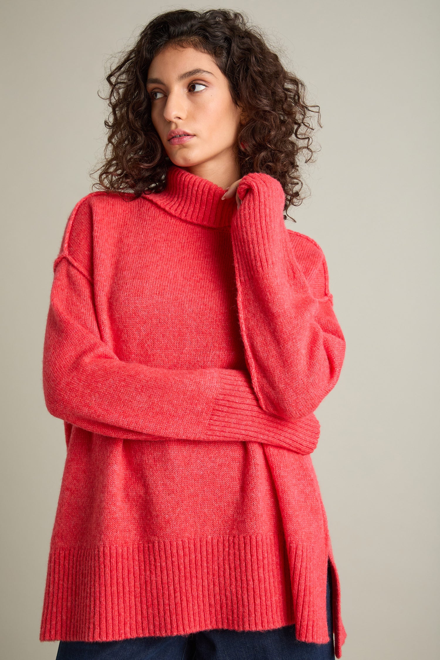 Person with curly hair wearing a bright red Charming Merino Polo Jumper with subtle cable knit detailing looks to their left against a plain background.