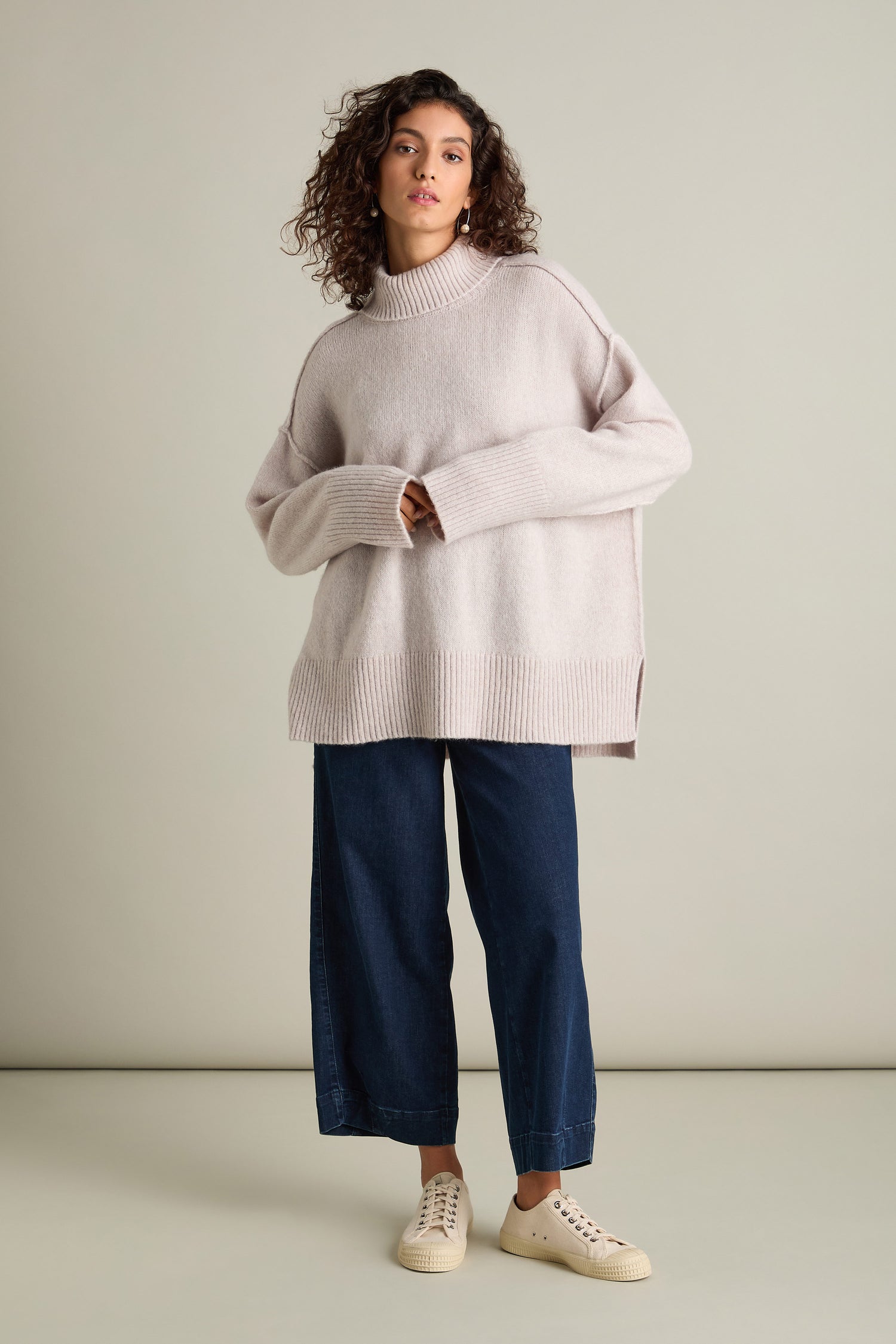 A person with curly hair wears a Charming Merino Polo Jumper, paired with wide-leg jeans and white sneakers, posing against a plain background.