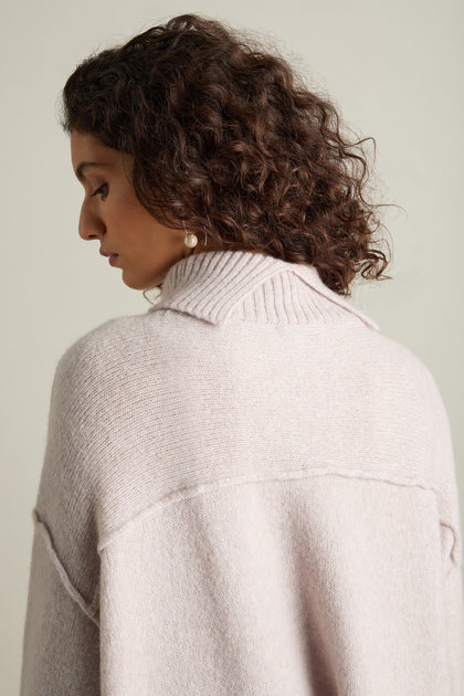 A woman with curly hair is displayed from the side and back, donning a light pink Charming Merino Polo Jumper. This refined sweater, crafted from extrafine merino wool, is elegantly designed with a collar and accompanied by pearl earrings for an added touch of sophistication.